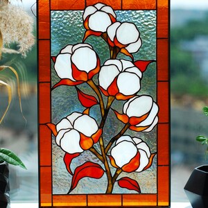 May include: Stained glass panel featuring a cotton plant design. The artwork showcases white cotton bolls with orange and red accents, set against a textured, light blue background. The panel is framed with an orange border.