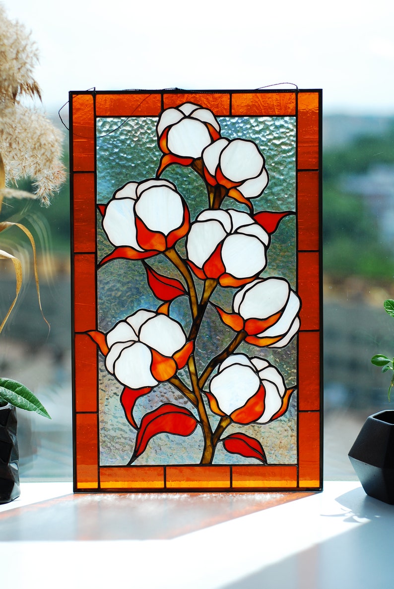 May include: Stained glass panel featuring a cotton plant design. The artwork showcases white cotton bolls with orange and red accents, set against a textured, light blue background. The panel is framed with an orange border.
