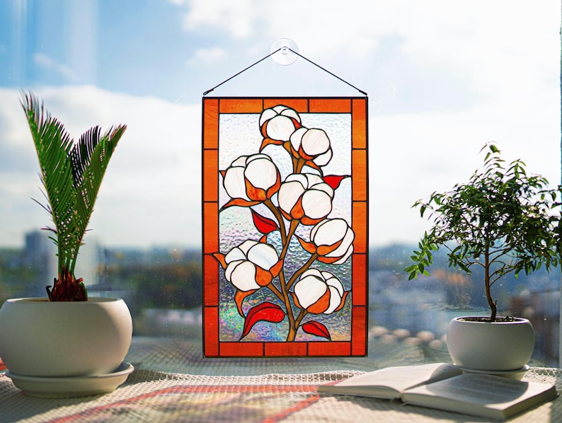 May include: Stained glass panel featuring a cotton plant design with white cotton bolls, orange accents, and red leaves. The panel is framed in orange and hangs in front of a window. Decorative art for home decor.
