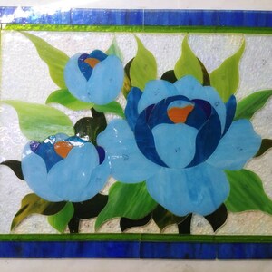 May include: A stained glass artwork featuring three blue flowers with green leaves. The flowers are arranged in a cluster and are surrounded by a green border. The artwork is set against a white background.