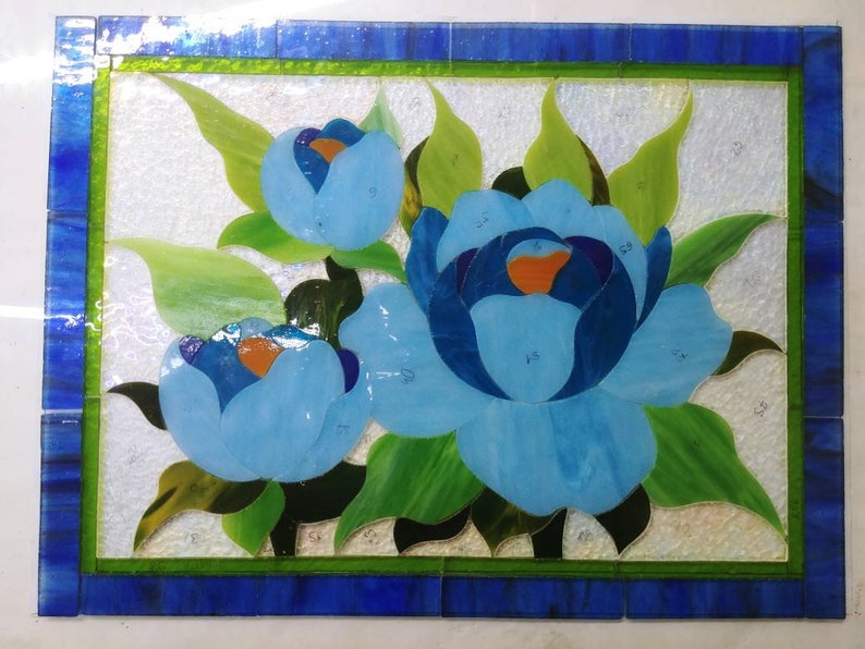 May include: A stained glass artwork featuring three blue flowers with green leaves. The flowers are arranged in a cluster and are surrounded by a green border. The artwork is set against a white background.