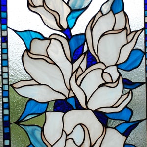 Puede incluir: Una ventana de vitrales que representa flores blancas de magnolia con hojas azules y verdes. Las flores est&aacute;n dispuestas en un racimo, con la flor m&aacute;s grande en el centro.