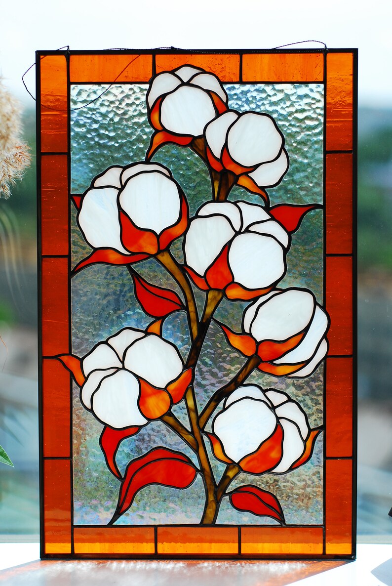May include: Stained glass panel featuring a cotton plant design. The artwork showcases white cotton bolls with orange and red accents, set against a textured glass background. The panel is framed with an orange border.