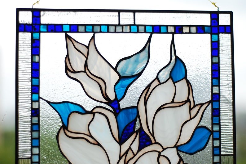 Puede incluir: Una ventana de vitrales que representa flores blancas con acentos azules y azul claro. Las flores est&aacute;n dispuestas en un racimo y est&aacute;n rodeadas por un borde azul oscuro.