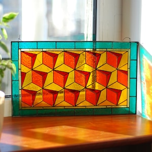 Op de afbeelding: Gebrandschilderd glasraam met een geometrisch patroon van rode en gele kubussen, omlijst door een turquoise rand. Het raam staat op een houten oppervlak, met een plant in een pot op de achtergrond.