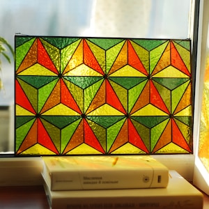 May include: A stained glass window panel with a geometric pattern of green, yellow, orange, and red triangles. The panel is framed in black and is illuminated by sunlight.