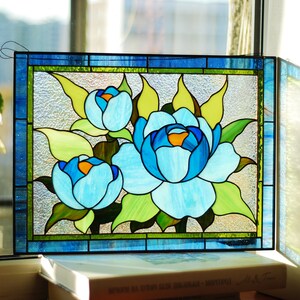 May include: A stained glass window panel depicting three blue peonies with green leaves. The panel is framed in blue and green glass.