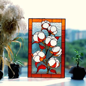 May include: Stained glass panel featuring a cotton plant with white and orange blossoms. The artwork is framed in a warm orange border. The panel is displayed on a windowsill with plants and a view of a cityscape.