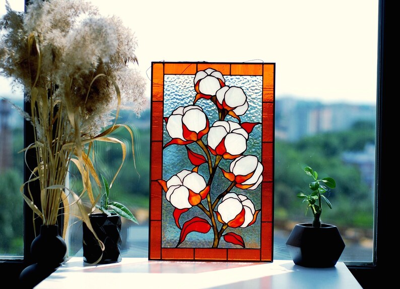 May include: Stained glass panel featuring a cotton plant with white and orange blossoms. The artwork is framed in a warm orange border. The panel is displayed on a windowsill with plants and a view of a cityscape.