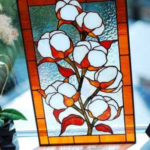 May include: Stained glass panel featuring a cotton plant design. The artwork showcases white cotton bolls with orange and red accents, set against a textured, light blue background. The panel is framed with an orange border.