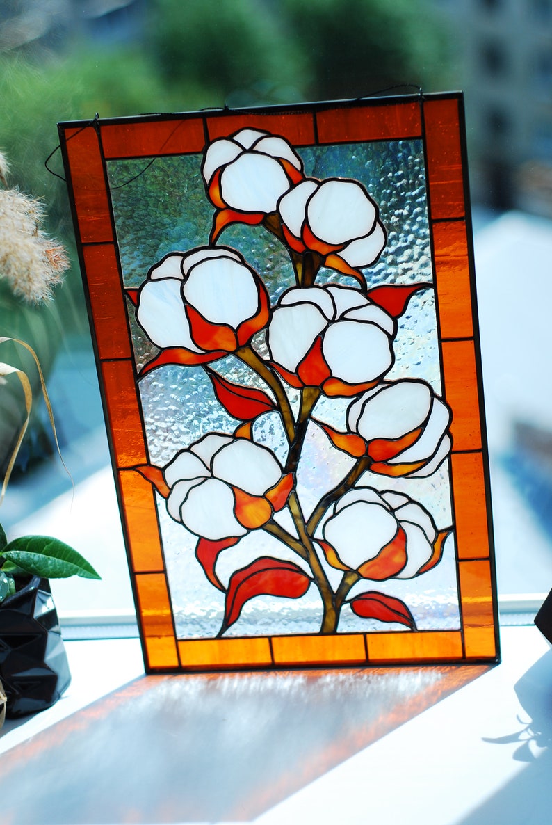 May include: Stained glass panel featuring a cotton plant design. The artwork showcases white cotton bolls with orange and red accents, set against a textured, light blue background. The panel is framed with an orange border.