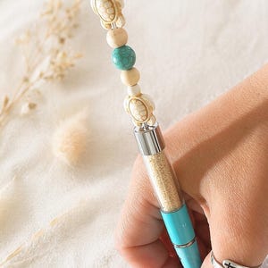 May include: A turquoise pen with a clear barrel filled with sand-like material. The pen's top features decorative beads, including turtle-shaped charms and turquoise and cream-colored beads. The pen is held in a hand, with a silver ring on the finger.