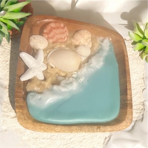 May include: A wooden tray with a beach scene. The tray contains a light blue resin ocean, tan sand, and various seashells and a white starfish. The tray is square with rounded corners.