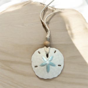 May include: A white ceramic sand dollar pendant with a blue and silver star design, hanging from a natural twine cord with a wooden bead.