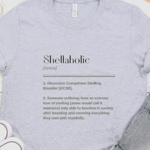 May include: A light gray t-shirt with the word "Shellaholic" printed in black, along with a definition. The definition describes "Obsessive Compulsive Shelling Disorder (OCSD)" and someone with an extreme love of shelling.