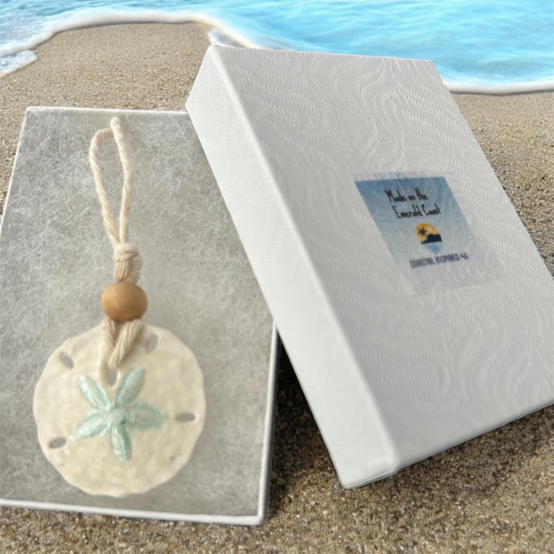 May include: A white sand dollar ornament with a light blue starfish design, hanging from a natural twine cord with a wooden bead. The ornament is packaged in a white box with the text "Made on the Emerald Coast" and "Destin, Florida 32541".