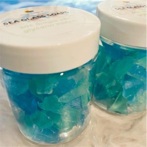 May include: Two clear glass jars filled with blue and green sea glass. The jars have white lids with labels that read "Sea Glass Soaps 80% Single Use Glycerin Soaps".
