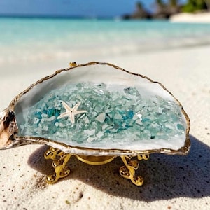 May include: An oyster shell dish with a gold-painted rim, filled with small, light blue and clear glass fragments and a single white starfish. The shell is supported by a decorative gold-colored metal stand with three ornate, curved legs.