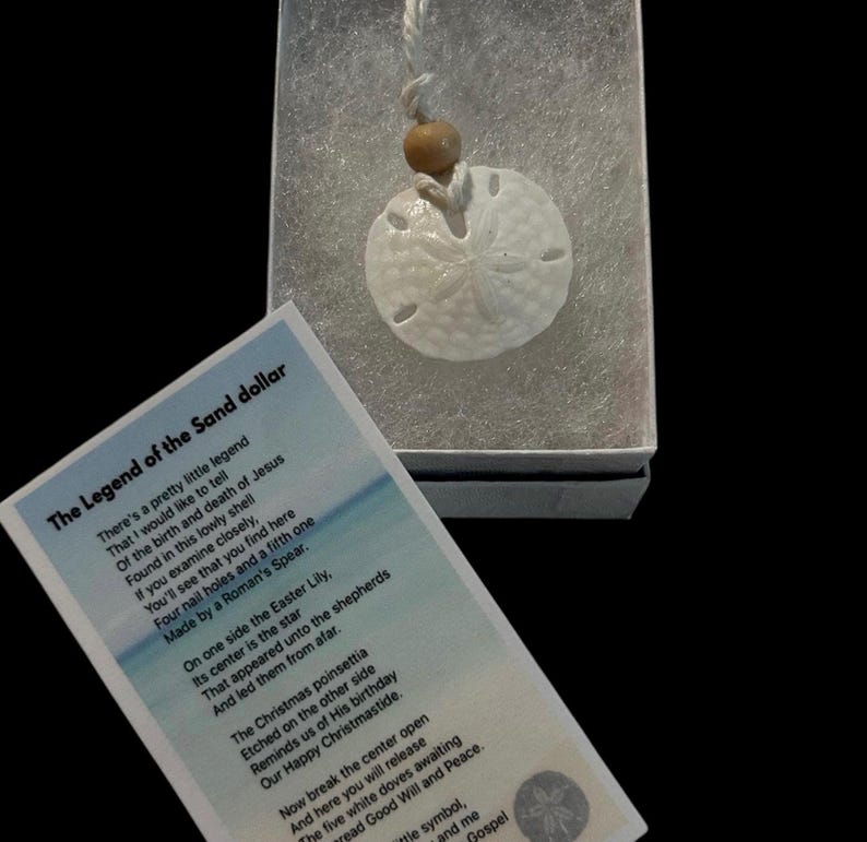May include: A white sand dollar necklace with a brown wooden bead and a card that reads "The Legend of the Sand Dollar" with a poem about the religious significance of the sand dollar.