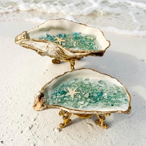 May include: Two decorative oyster shells with gold-painted edges, each resting on a small gold metal stand. The interior of each shell is filled with crushed blue and green glass fragments and features a single small white starfish accent.