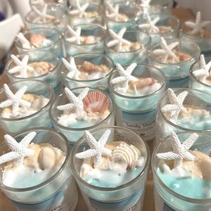 May include: A collection of small glass candles with a beach theme. Each candle features a light blue wax base, topped with white sand, seashells, and a white starfish. The candles are arranged in a grid pattern.