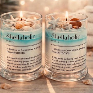 May include: Two lit glass jar candles with a beach theme. Each candle has a blue and white wax design, topped with seashells and starfish. The text "Shellaholic" is printed on the front.