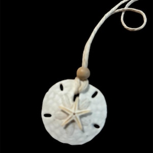 May include: A white sand dollar necklace with a starfish charm and a wooden bead. The necklace is made of white cord.
