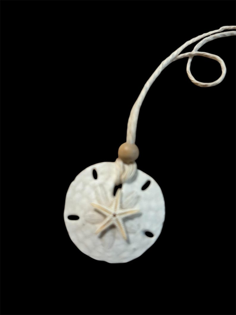 May include: A white sand dollar necklace with a starfish charm and a wooden bead. The necklace is made of white cord.