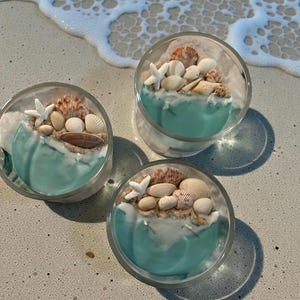 May include: Three clear glass candles with a turquoise wave design, topped with seashells, starfish, and sand. The candles are arranged on a sandy surface near the ocean's edge, with foamy waves in the background. The candles are a decorative item.