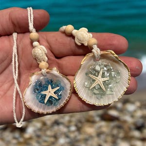 May include: Two seashell pendants with gold trim, each filled with blue or green glass and a starfish. The pendants are strung on a white cord with wooden beads and turtle charms, perfect for beach-themed decor.