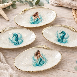 May include: Four oval-shaped white shell dishes with gold-painted edges. Two dishes feature a central illustration of a mermaid emerging from blue water, and two dishes feature a central illustration of a blue seahorse surrounded by water splashes.