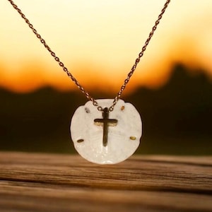 May include: A gold-toned necklace featuring a white sand dollar pendant with a gold cross charm. The necklace hangs against a blurred background of warm sunset colors, resting on a wooden surface.