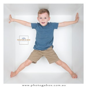 Może przedstawiać: Młody chłopiec w niebieskiej, pasiastych koszulach i krótkich spodenkach khaki stoi w białym pudełku. Trzyma się boków pudełka rękami, a nogi ma szeroko rozstawione. Chłopiec uśmiecha się i patrzy w obiektyw. Tekst "PHOTOG-A-BOX PHOTOGRAPHY" jest widoczny w lewym dolnym rogu obrazu.