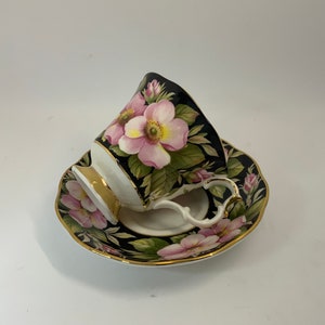 May include: A black teacup and saucer with a floral design. The cup and saucer are decorated with pink and white roses on a black background.