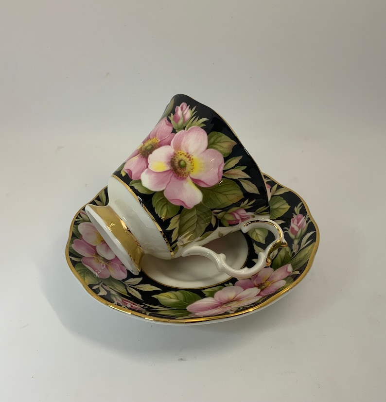 May include: A black teacup and saucer with a floral design. The cup and saucer are decorated with pink and white roses on a black background.