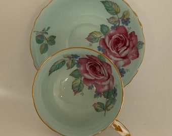 May include: A light blue teacup and saucer set with a floral design featuring pink roses and green leaves. The set has a gold trim around the edges.