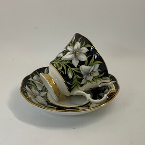 May include: A black teacup and saucer with a white floral design. The cup and saucer have a gold trim.