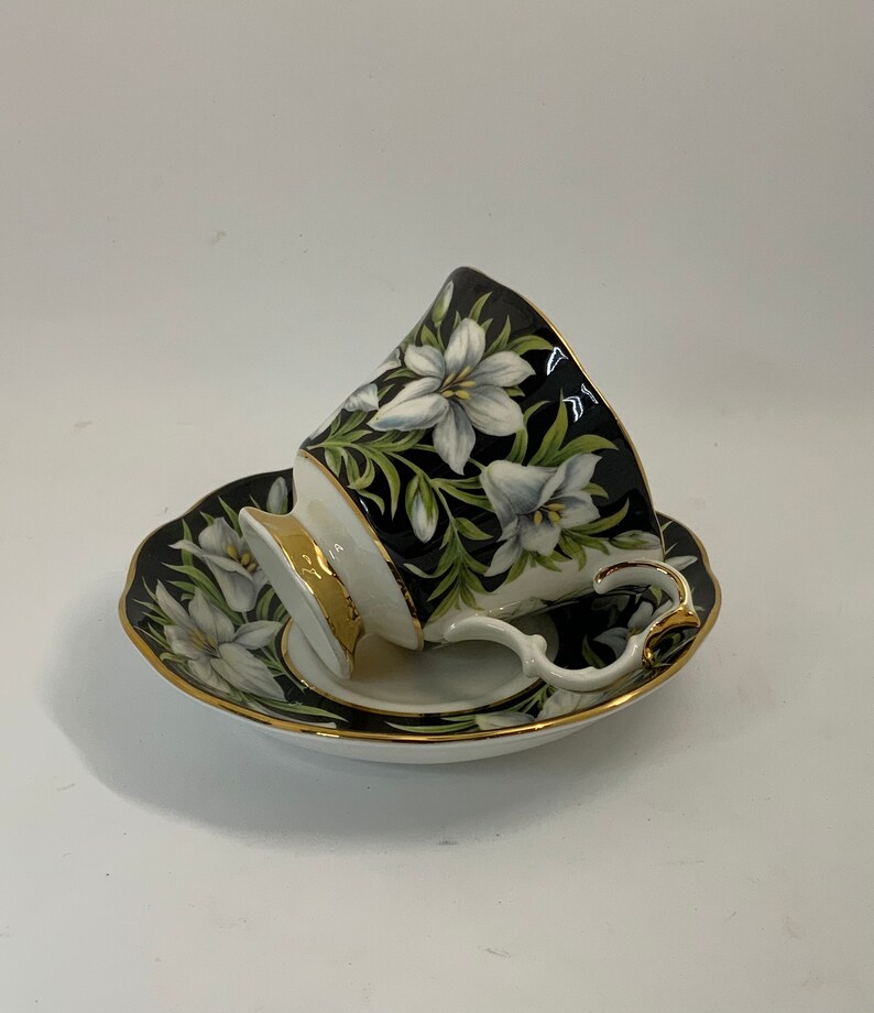 May include: A black teacup and saucer with a white floral design. The cup and saucer have a gold trim.