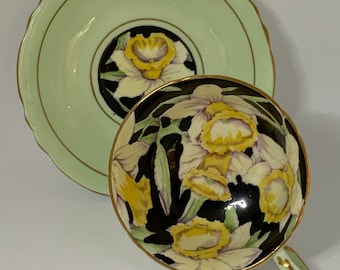 May include: A vintage teacup and saucer set with a black background and yellow daffodil floral pattern. The saucer is a pale green color with a gold rim.