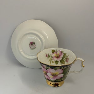 May include: A white teacup and saucer set with a black and gold floral design. The cup features pink roses and green leaves on a black background with a gold rim. The saucer is white with a gold rim.