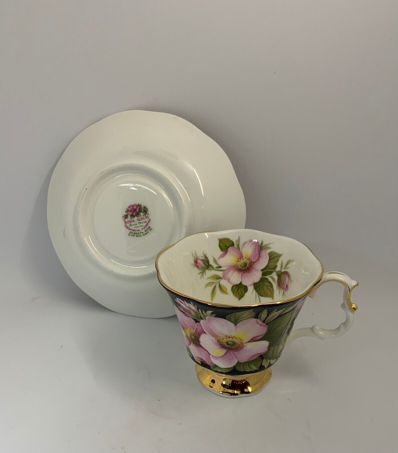 May include: A white teacup and saucer set with a black and gold floral design. The cup features pink roses and green leaves on a black background with a gold rim. The saucer is white with a gold rim.