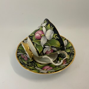 May include: A black and white teacup and saucer set with a floral design. The cup and saucer are decorated with pink and white flowers and green leaves. The cup has a gold rim and the saucer has a gold trim.
