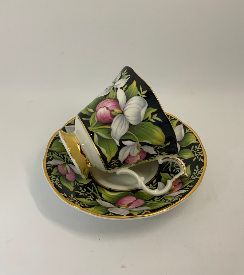 May include: A black and white teacup and saucer set with a floral design. The cup and saucer are decorated with pink and white flowers and green leaves. The cup has a gold rim and the saucer has a gold trim.