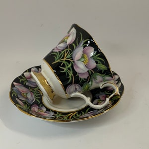 May include: A black teacup and saucer with a floral design. The cup and saucer are decorated with pink and white flowers on a black background. The cup has a gold rim and handle.