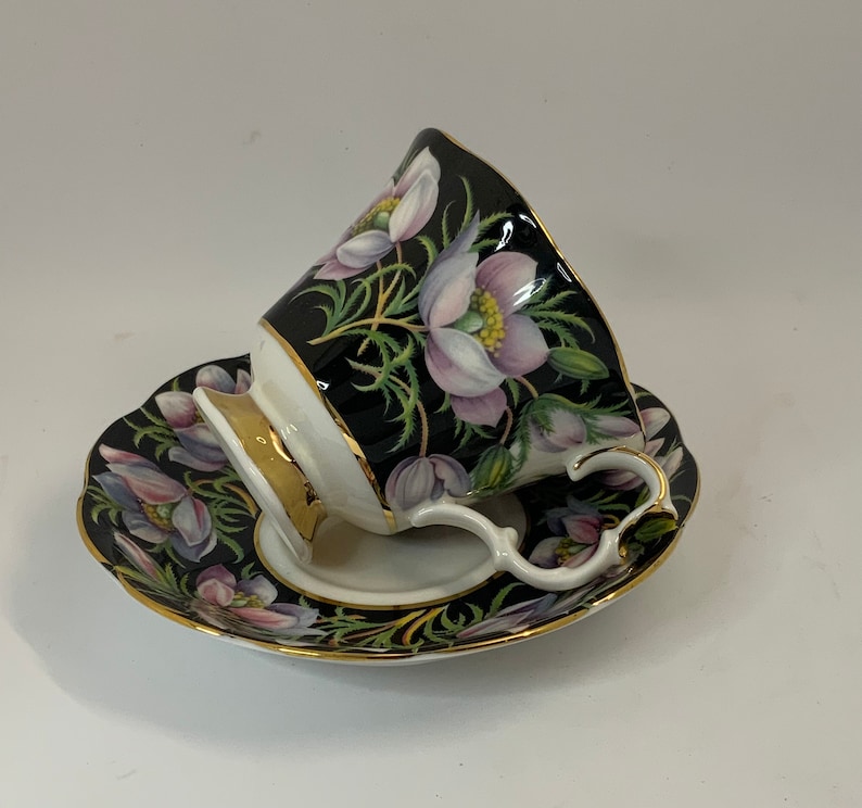 May include: A black teacup and saucer with a floral design. The cup and saucer are decorated with pink and white flowers on a black background. The cup has a gold rim and handle.
