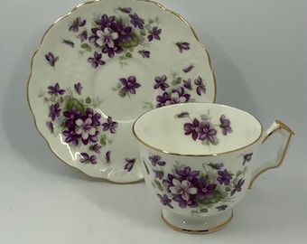 Può includere: Una tazzina da tè e un piattino bianchi con un delicato motivo floreale con violette viola e bianche. Il set ha un bordo dorato.