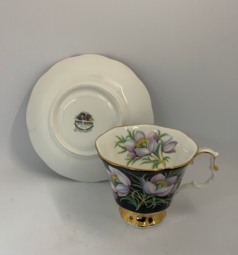 May include: A white teacup and saucer with a black and gold floral design. The saucer has a gold rim and the teacup has a gold handle. The teacup features a floral pattern with white flowers and green leaves on a black background. The saucer has the Royal Albert logo and the words "Botanical Flowers" in gold lettering.