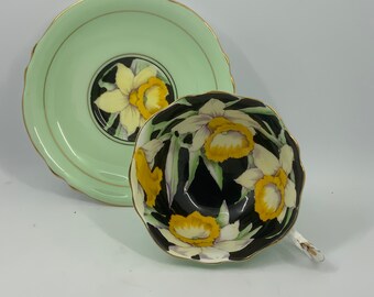 May include: A vintage teacup and saucer set with a black background and yellow and white daffodil floral pattern. The saucer is a light green color with a gold trim.