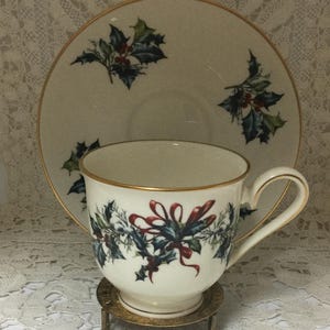 May include: A white teacup and saucer with a gold rim and a holly and ribbon design. The cup and saucer are on a gold metal stand.