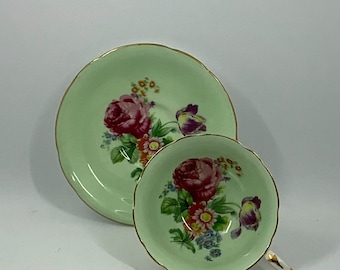 May include: A vintage teacup and saucer set with a delicate floral design. The cup and saucer are a pale green color with a gold rim. The floral design features pink roses, purple tulips, and other colorful flowers.